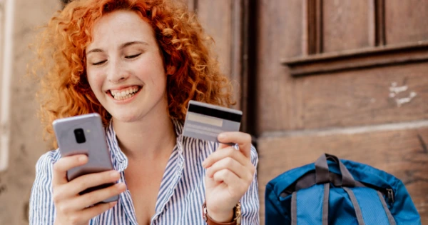 A woman smiling while looking at her phone and holding a credit card.