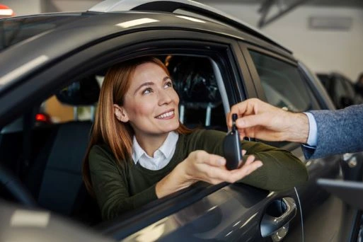 A woman sitting in a car smiles as someone hands her the keys.